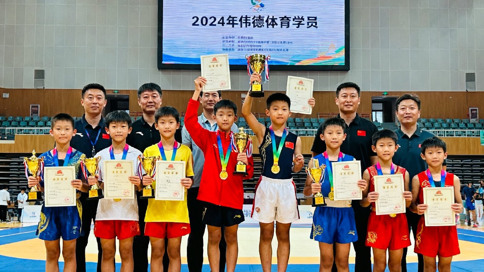 2024年伟德体育学员在多项省市比赛中获奖后的集体合影,十几位青少年选手穿着各色奖牌站在领奖台前,有的手持奖杯,有的举着证书,教练团队站在后排,背景是比赛场馆的巨大电子屏幕。